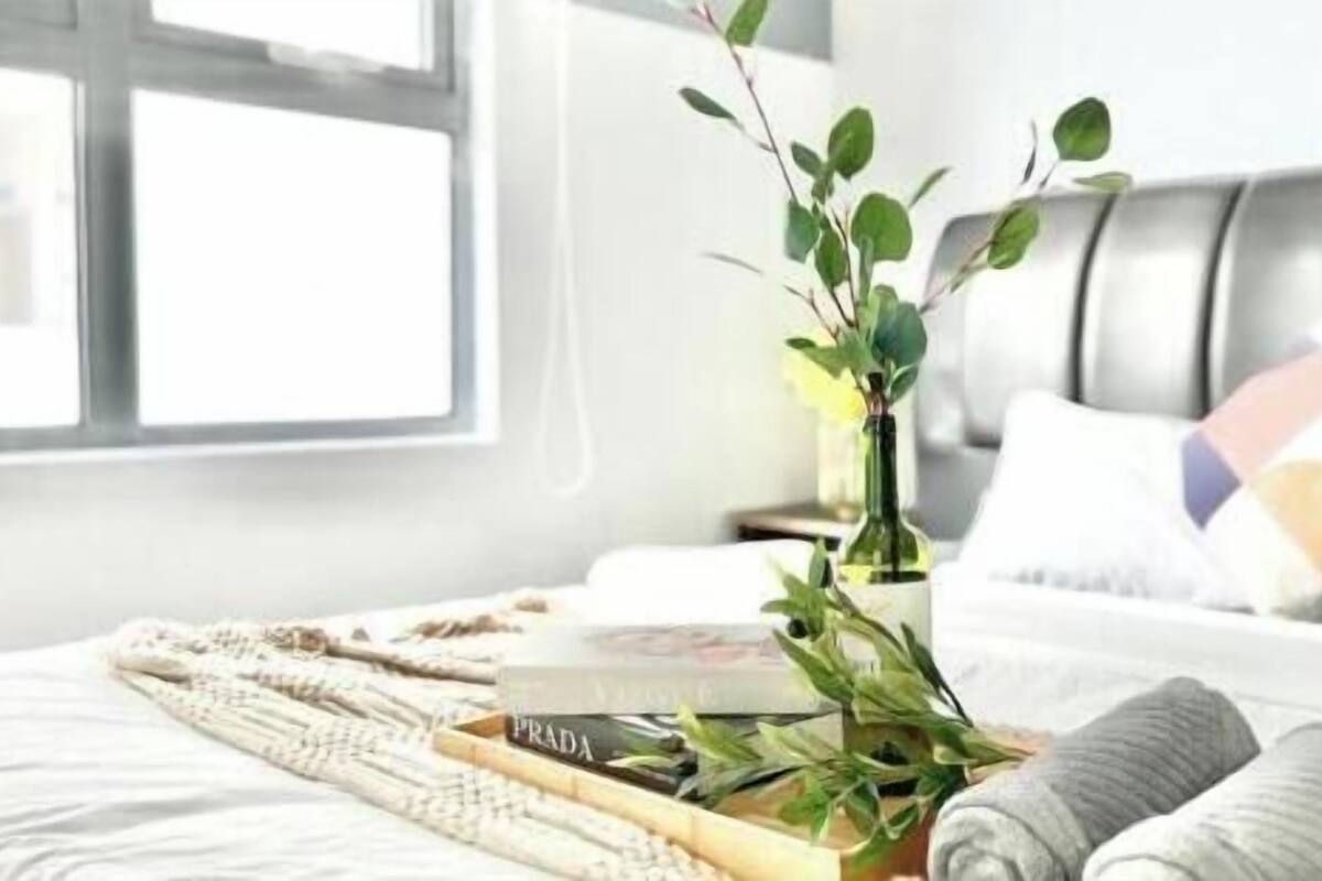Een netjes opgemaakt bed met een witte deken, een opgerolde handdoek, een dienblad met boeken en een potplant.