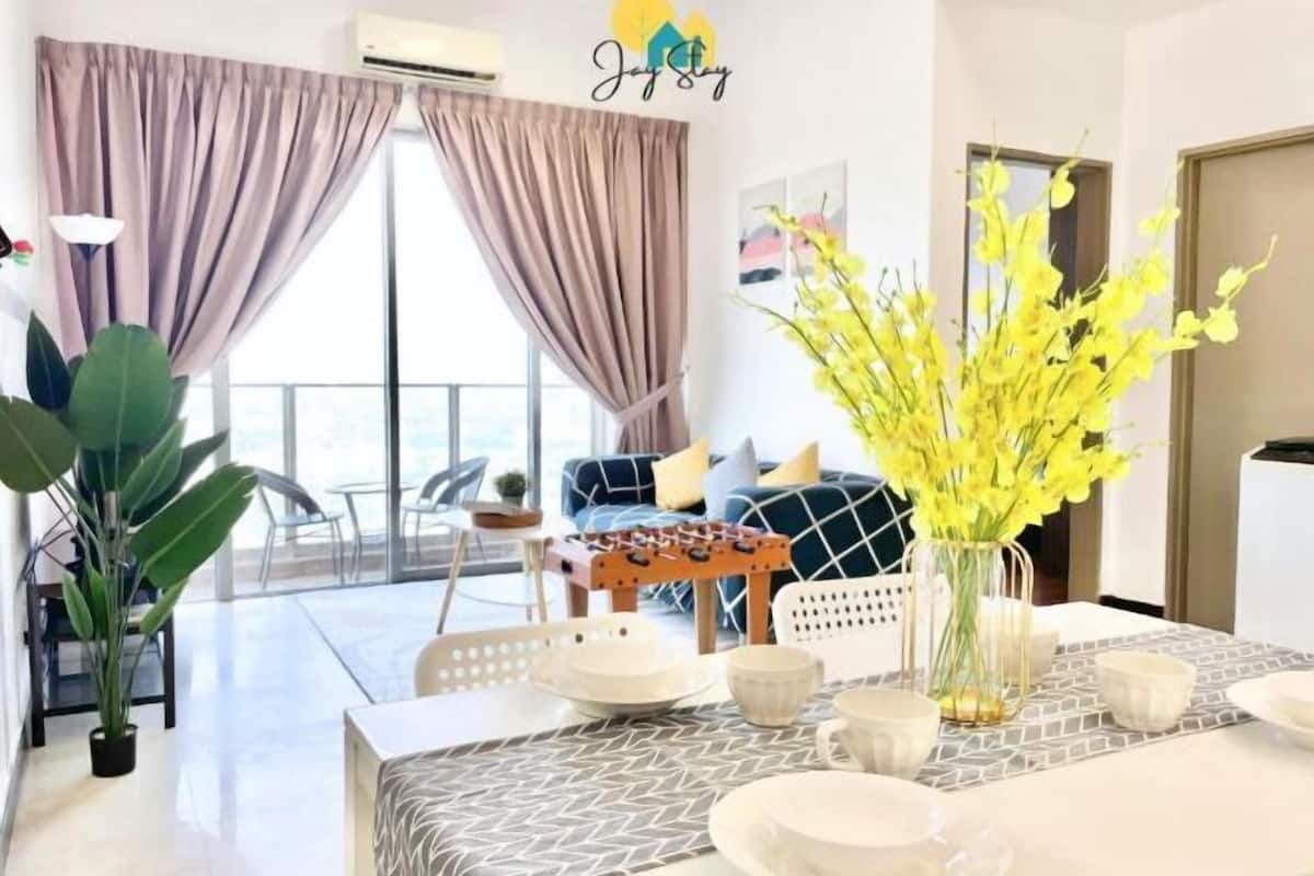 Een moderne woonkamer met een eethoek, grote ramen voorzien van gordijnen, een bank met kussens, een salontafel en een vaas met gele bloemen.