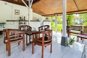 Un espacio de comedor al aire libre con techo, mesas y sillas de madera, una planta en maceta y una casa al fondo.