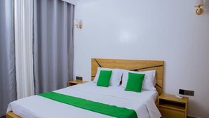 A neatly made bed with a wooden headboard, white bedding, and green pillows. There are two bedside tables with lamps, a window with curtains, and a wall-mounted light fixture. - Kigali Bliss Apartment (Kigali)