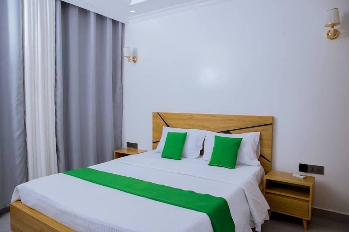 A neatly made bed with a wooden headboard, white bedding, and green pillows. There are two bedside tables with lamps, a window with curtains, and a wall-mounted light fixture.