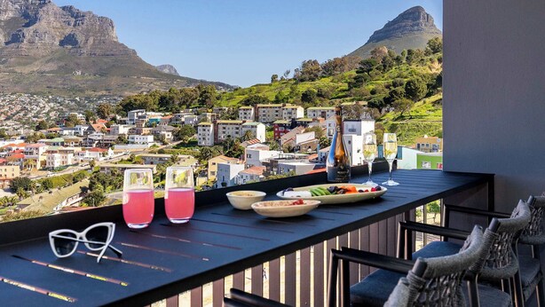 Ein Balkon mit Esstisch, Gläsern und einer Flasche, mit Blick auf eine Stadt mit Gebäuden und Bergen.