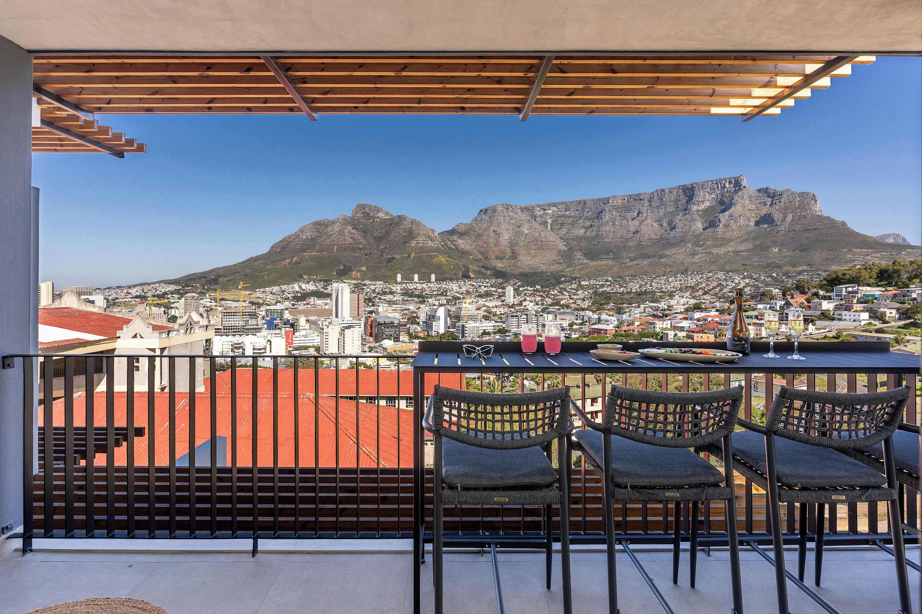 A balcony with chairs and a table, overlooking a cityscape and a mountain.
