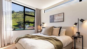 Ein Schlafzimmer mit einem großen Fenster, einem Bett, Nachttischlampen, einem gerahmten Bild und einem Nachttisch.