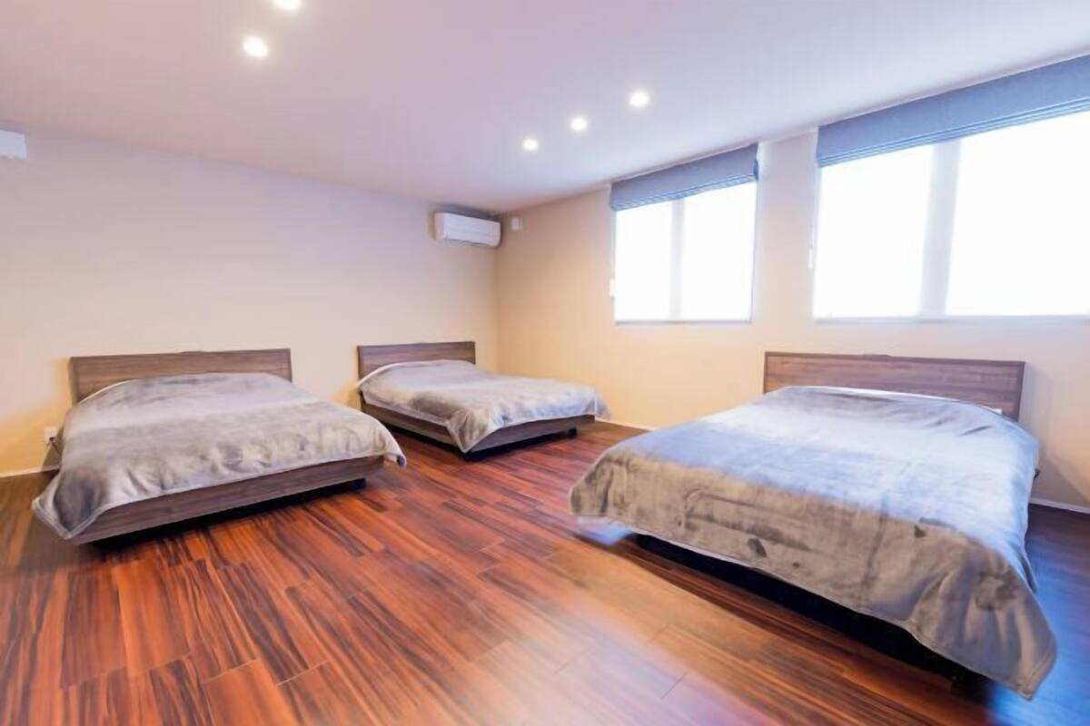 A room with three single beds, wooden flooring, and a window with blinds.