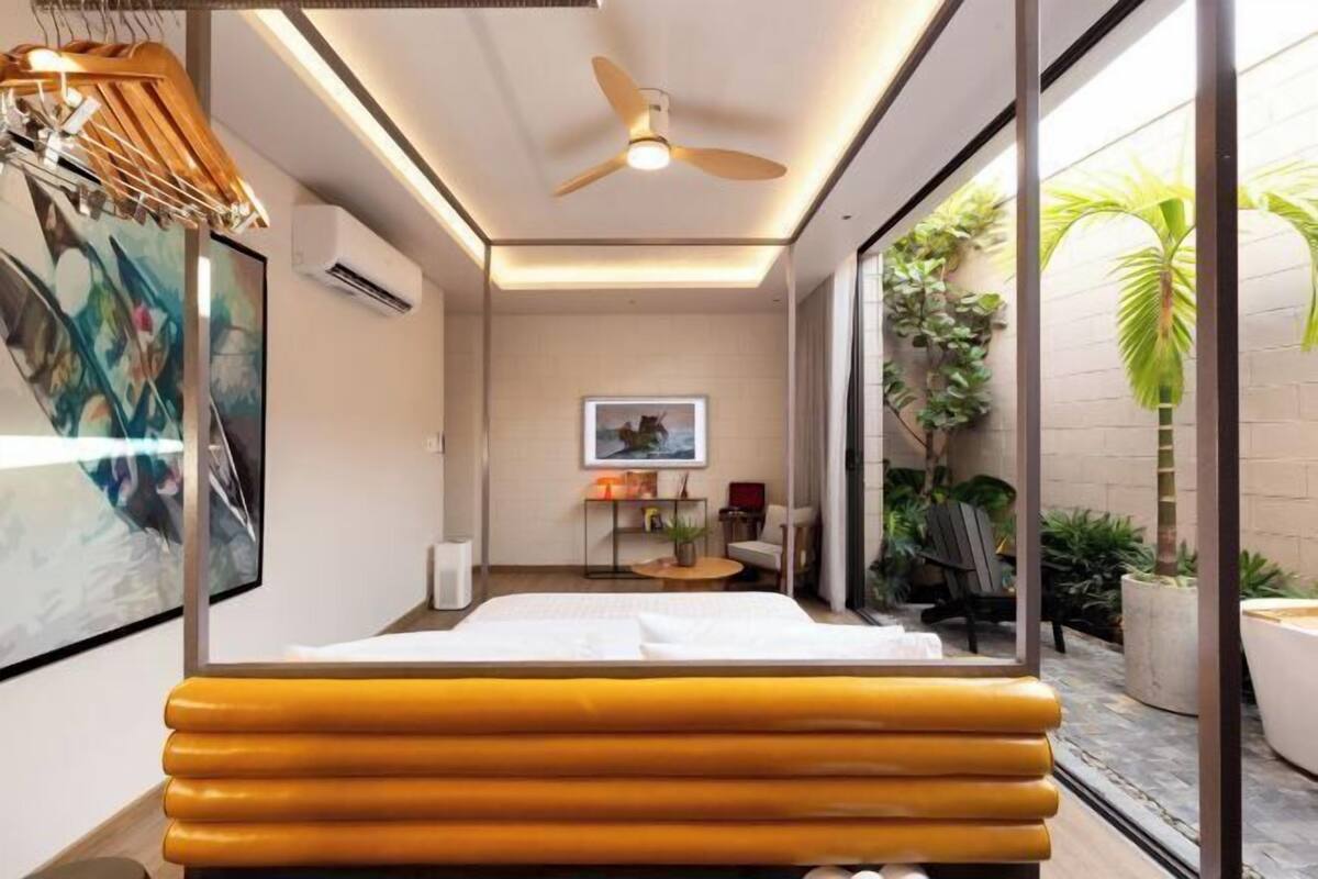 A modern hotel room with a bed, a ceiling fan, a window with a view of the garden, and a painting on the wall.
