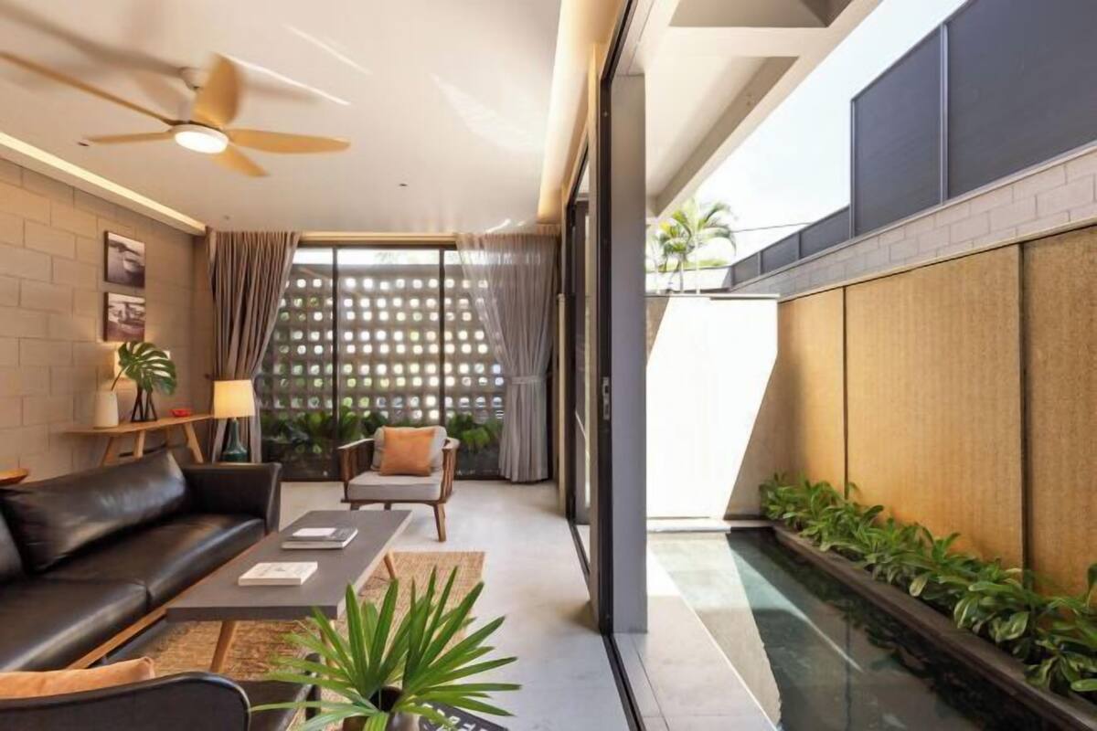 A modern living room with a sofa, coffee table, and a ceiling fan. There is a view of a balcony with plants and a lattice fence.