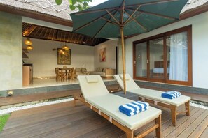 Una terraza de madera con dos sillas de descanso y una mesa, un techo de paja y un ventanal grande.