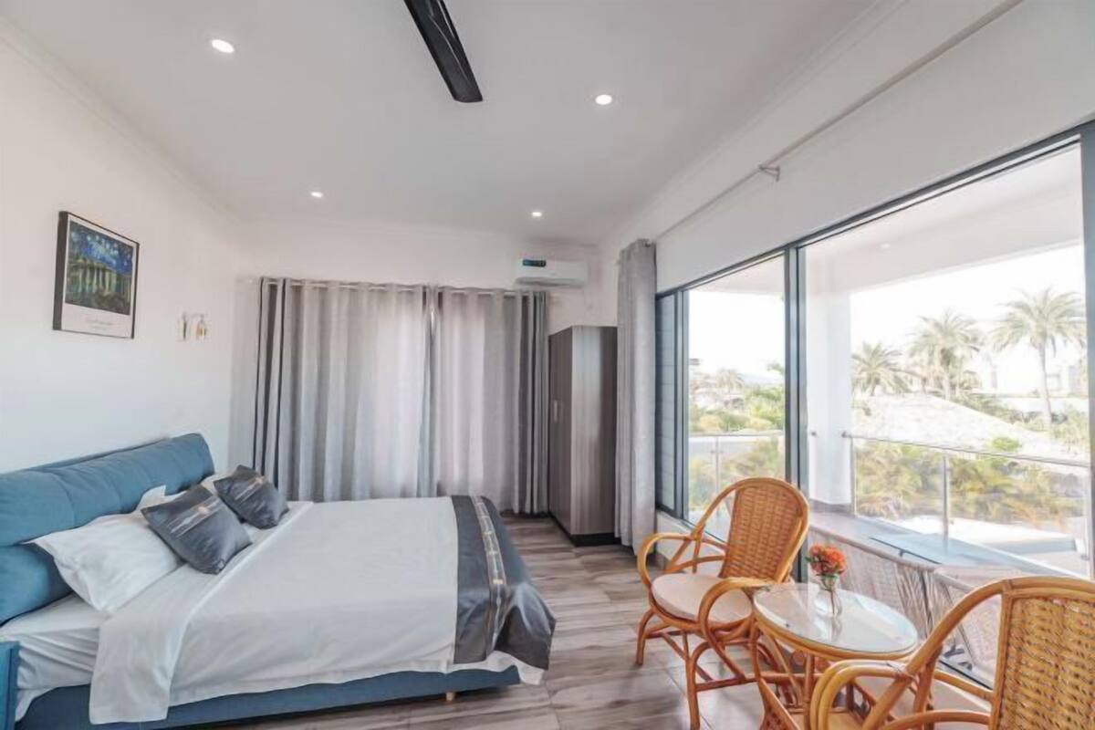 Quarto de hotel moderno com uma cama, varanda com cadeiras de vime e uma janela grande com vista.