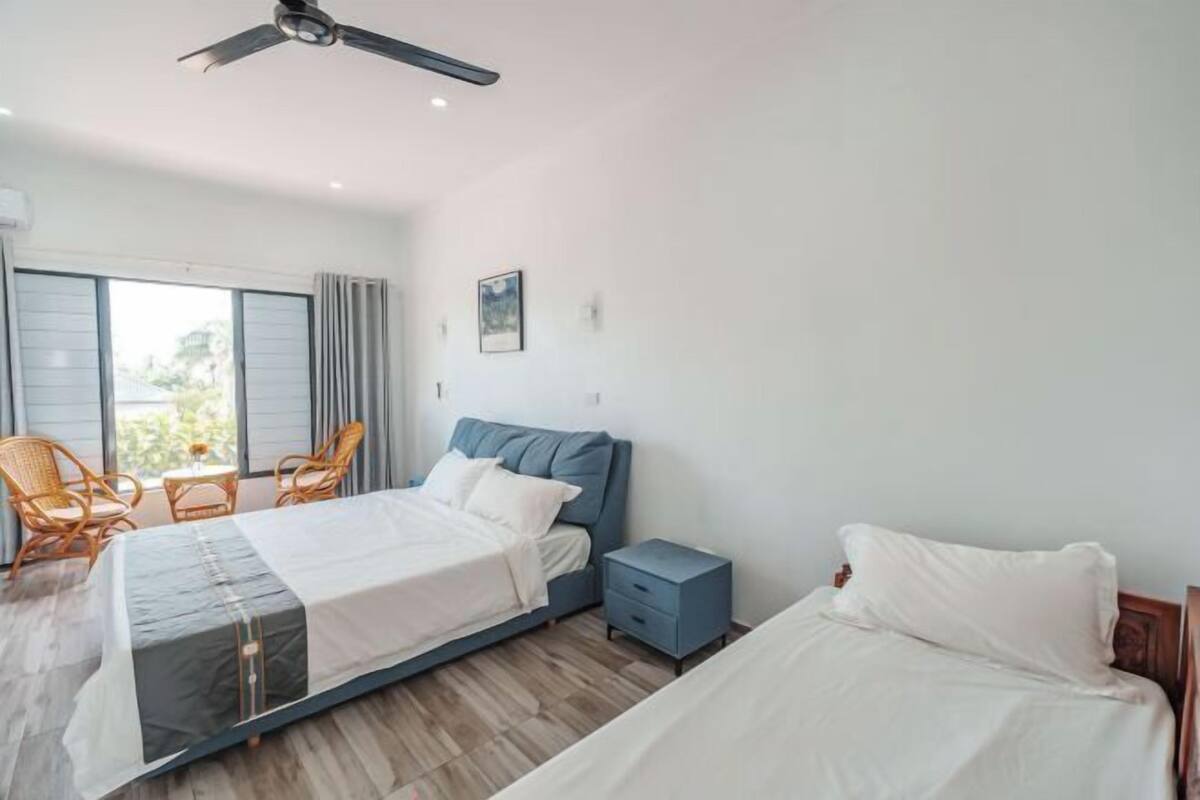 Quarto com cama, mesa de cabeceira, cadeira, ventoinha no teto e janela.