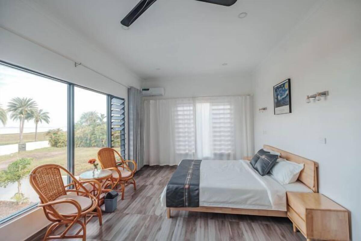 Quarto com cama, cadeira, mesa e uma janela grande com vista para palmeiras.