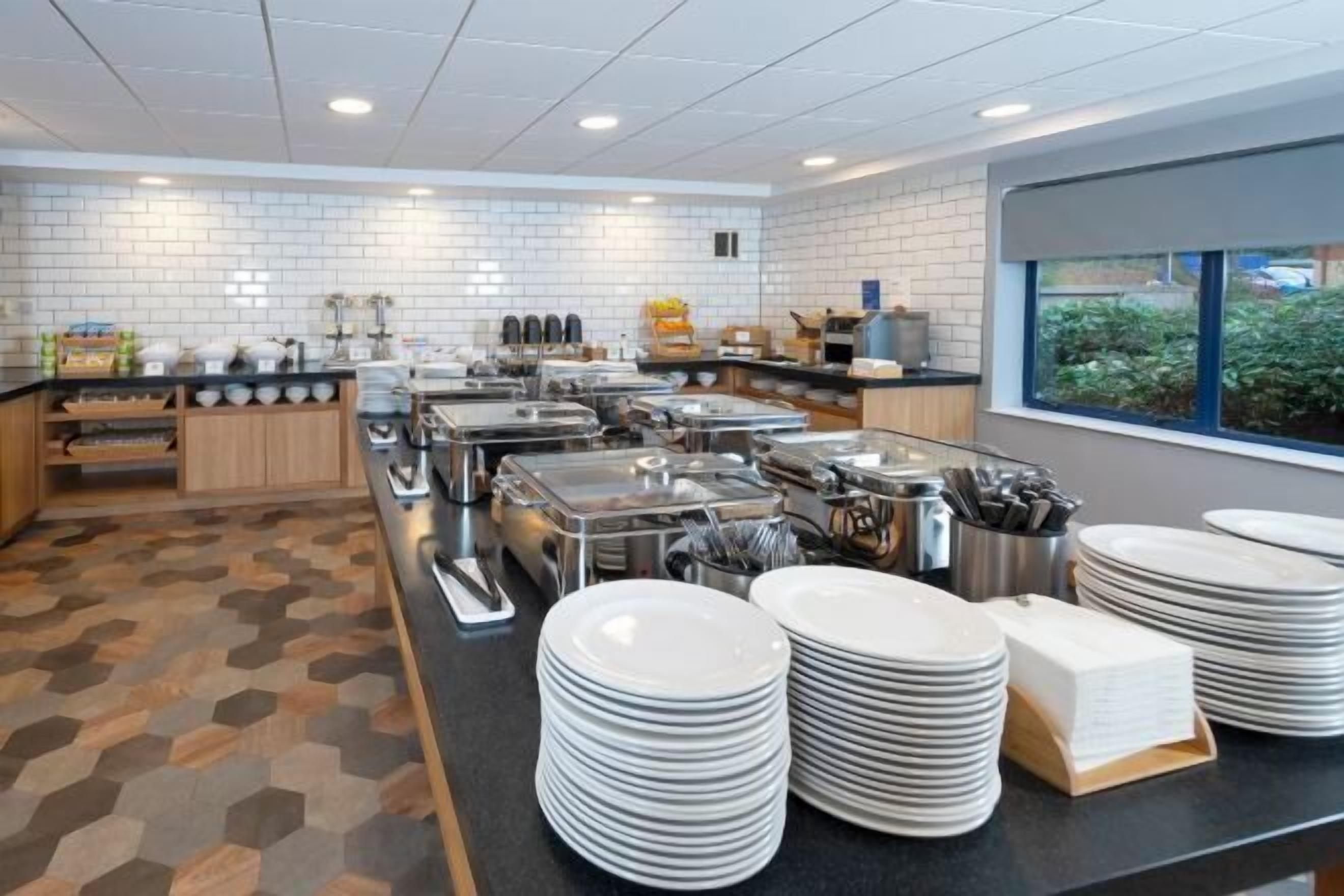 En buffetopstilling med flere serveringsstationer, et flisebeklædt bagpanel og et vindue med udsigt til grønne omgivelser.