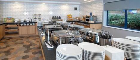 En buffetopstilling med flere serveringsstationer, et flisebeklædt bagpanel og et vindue med udsigt til grønne omgivelser.