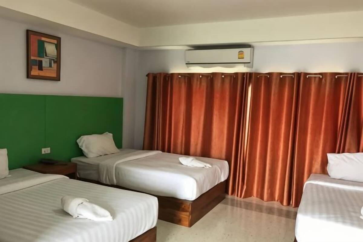 A hotel room with three beds, a green headboard, an air conditioning unit, and a window with brown curtains.