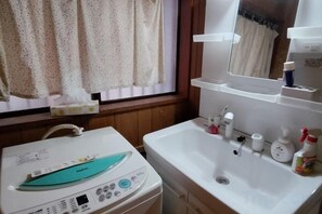 A bathroom with a washing machine, a sink with toiletries, and a window with curtains. - Cherry Blossom House (Saitama)