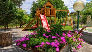 Un’area giochi con uno scivolo e strutture per arrampicata, circondata da un giardino fiorito di fiori rosa e una casetta di legno.