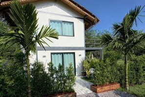 Une maison moderne à deux étages, dotée d’un balcon, entourée d’une végétation luxuriante et de palmiers.
