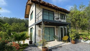 Ein zweistöckiges Gebäude mit einem Strohdach, einem Balkon mit Sitzgelegenheit und einem Garten mit Topfpflanzen und Blumen.