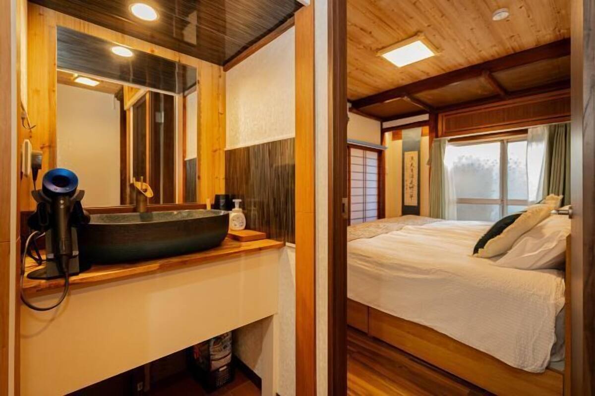Una habitación con techo de madera, cama con ropa blanca, un ventanal con cortinas y un lavabo con lavamanos.