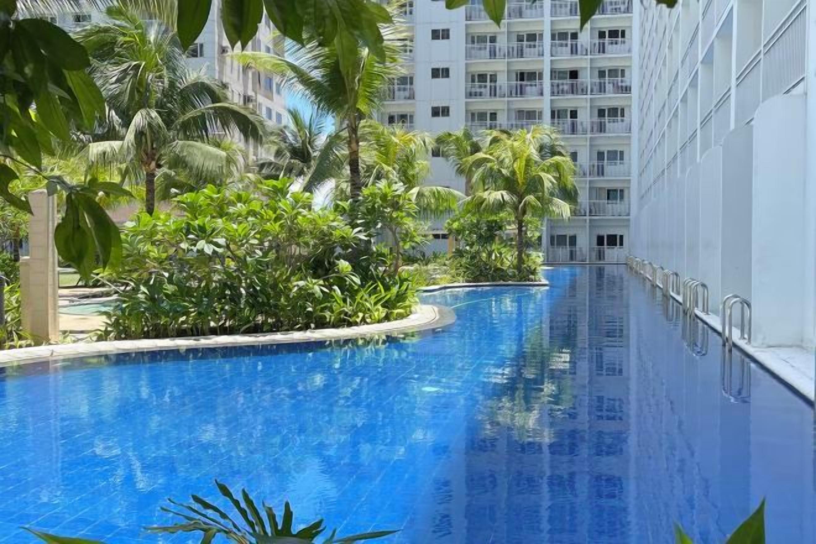 Une piscine à bord incurvé, entourée de verdure, avec un immeuble à plusieurs étages en arrière-plan.