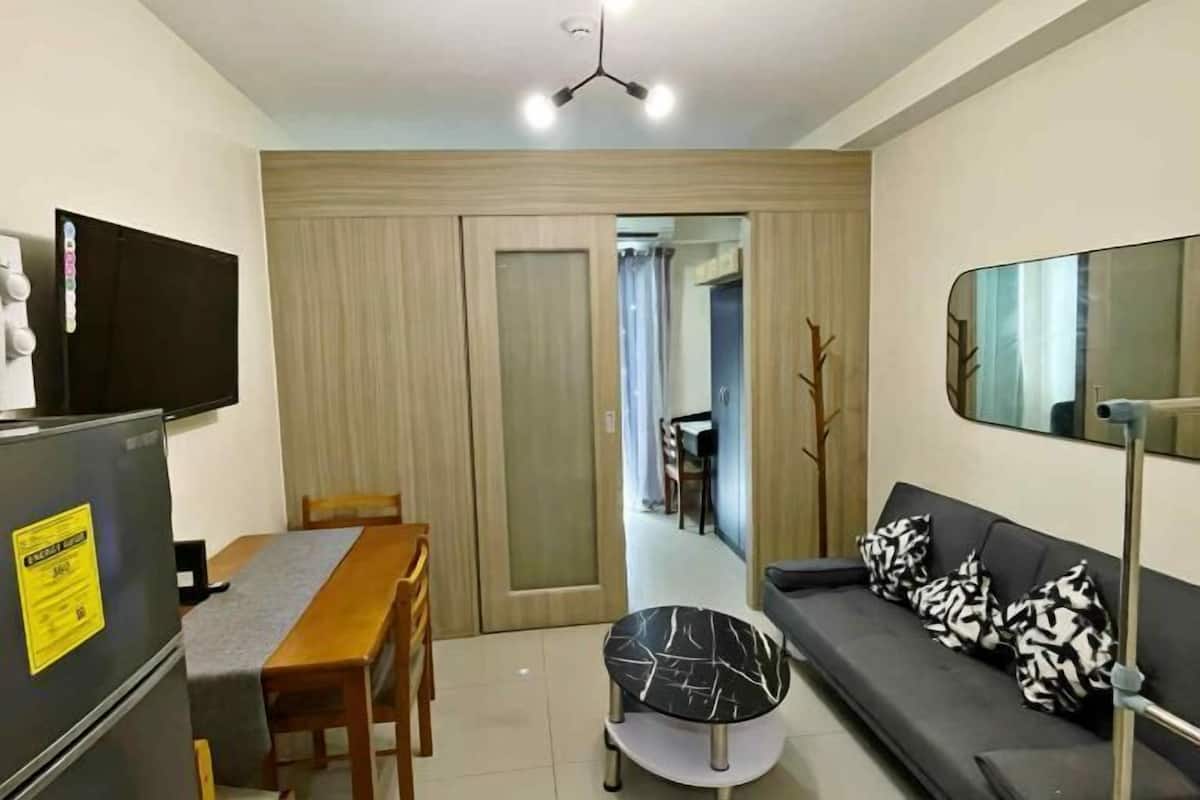 A compact living space with a dining table, a sofa with patterned cushions, a small round table, a refrigerator, and a television.