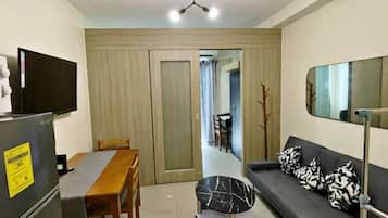 A compact living space with a dining table, a sofa with patterned cushions, a small round table, a refrigerator, and a television.