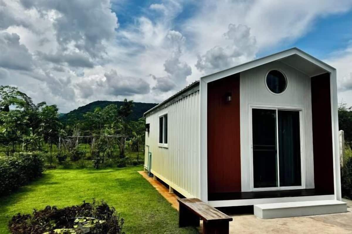Una pequeña casa de un piso con fachada blanca y puerta marrón, rodeada de un exuberante jardín verde y colinas al fondo.