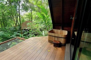 Une terrasse en bois avec une grande baignoire en bois et un petit tabouret, le tout entouré d’une végétation luxuriante.