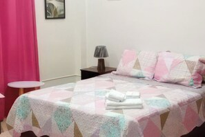 Una cama bien tendida con un colcha, una lámpara de noche y una mesita redonda con una cortina rosa al fondo.