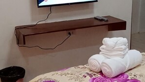 A hotel room with a bed, a TV mounted on the wall, a wooden shelf, and a trash bin. - Sunnyside Hotel (Santo Tomas)