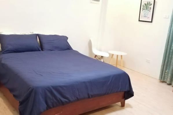 A bed with a blue cover, a white chair, and a small table with a plant on the wall.