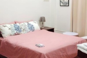 Una cama bien hecha con colcha rosa y almohadones florales, una lámpara de noche y un cuadro en la pared.