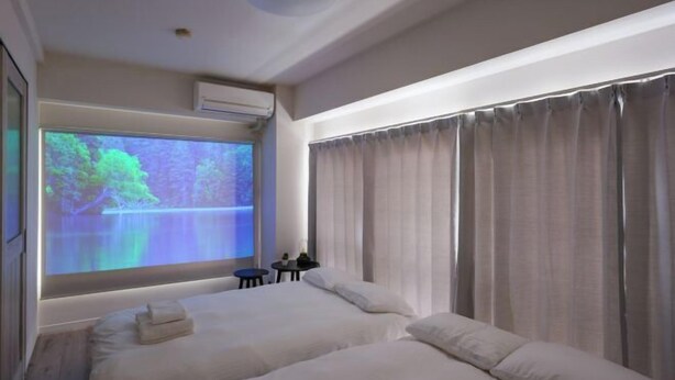 A hotel room with two beds, a large window with curtains, a projector screen displaying a nature scene, and a small table with a bottle. - Oyama Pantheon (Tokyo)