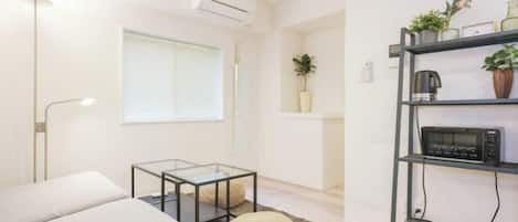 Una sala de estar moderna con un sofá, una mesa de centro de vidrio, un reposapiés redondo, una estantería con plantas y una lámpara de techo.