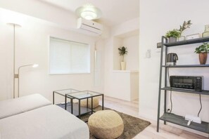 A modern living room with a sofa, a glass coffee table, a round ottoman, a bookshelf with plants, and a ceiling light fixture. - Oyama Pantheon (Tokyo)