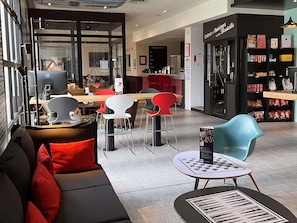 A modern cafe with a black sofa, red chairs, and a blue chair. There is a counter with a menu and a computer. - Ibis Villepinte Parc Expos (Villepinte)