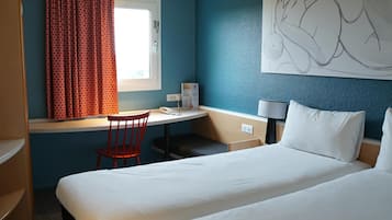 A hotel room with a bed, a desk with a chair, a window with curtains, a wall-mounted lamp, and a painting.