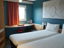 Una habitación de hotel con cama, escritorio y silla, ventana con cortinas, lámpara de pared y un cuadro.