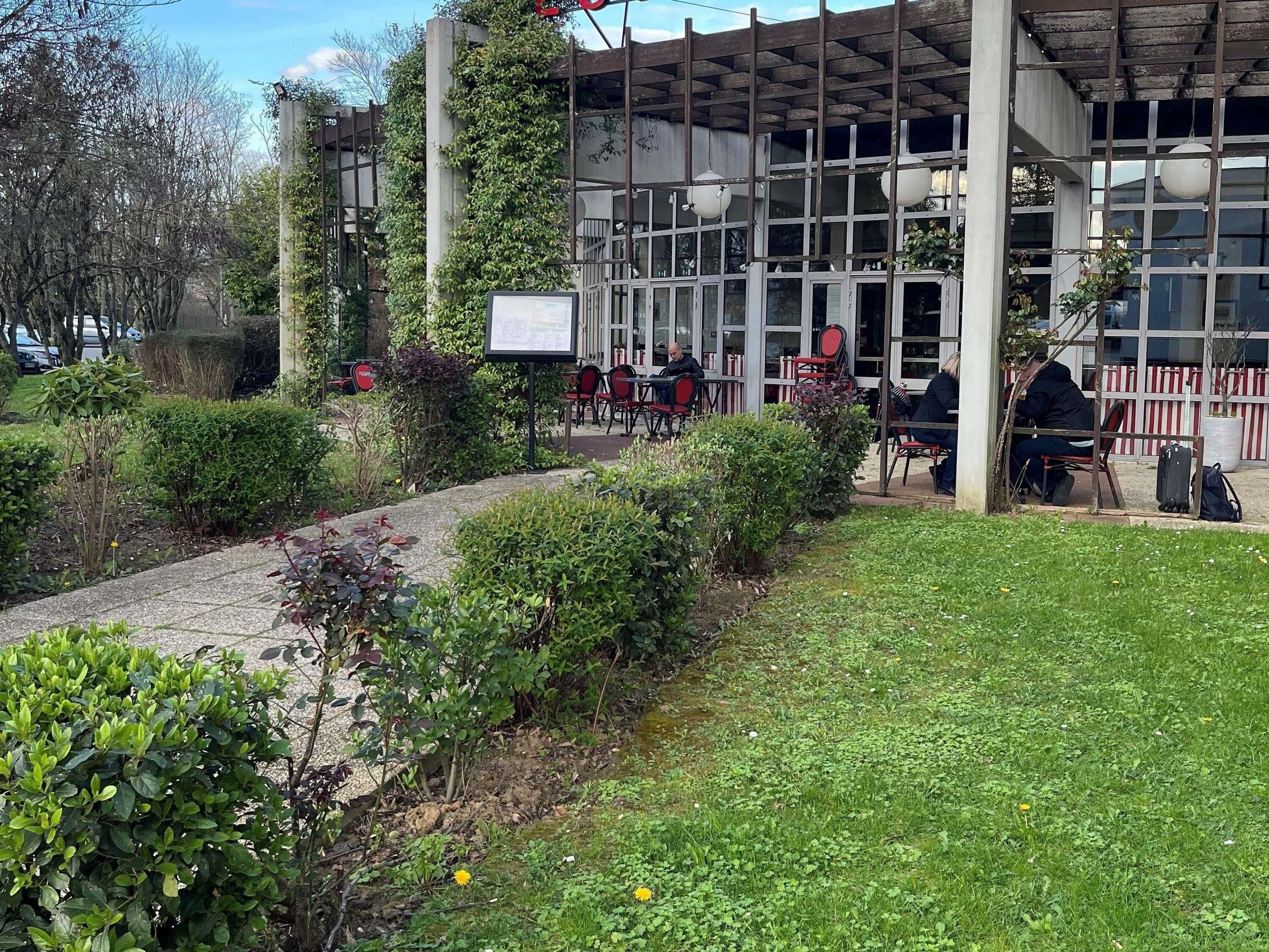 A building with a covered outdoor seating area, a paved walkway, and greenery.
