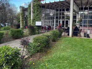 A building with a covered outdoor seating area, a paved walkway, and greenery. - Ibis Villepinte Parc Expos (Villepinte)