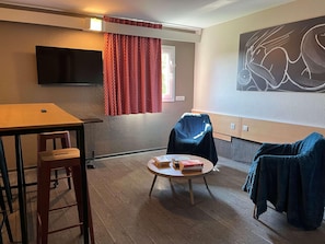 A room with a flat-screen TV, a round table with books, a chair, and a couch. - Ibis Villepinte Parc Expos (Villepinte)
