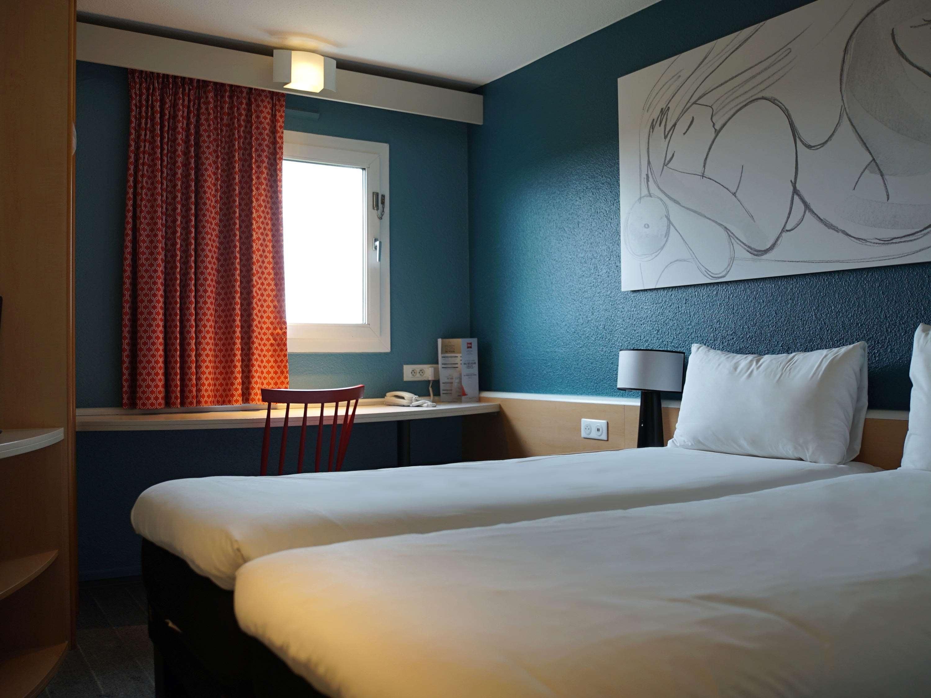 A hotel room with two beds, a desk with a chair, a window with curtains, a wall-mounted lamp, and a large abstract painting.