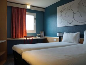 A hotel room with two beds, a desk with a chair, a window with curtains, a wall-mounted lamp, and a large abstract painting. - Ibis Villepinte Parc Expos (Villepinte)