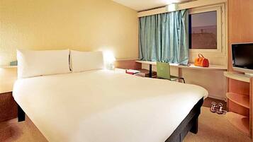 A hotel room with a large bed, a television, a window with curtains, and a small table with a bag and a pair of shoes.