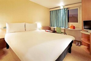 A hotel room with a large bed, a television, a window with curtains, and a small table with a bag and a pair of shoes. - Ibis Villepinte Parc Expos (Villepinte)