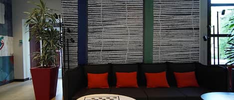 A modern lounge area with a black sofa, red cushions, a chess table, and a potted plant.