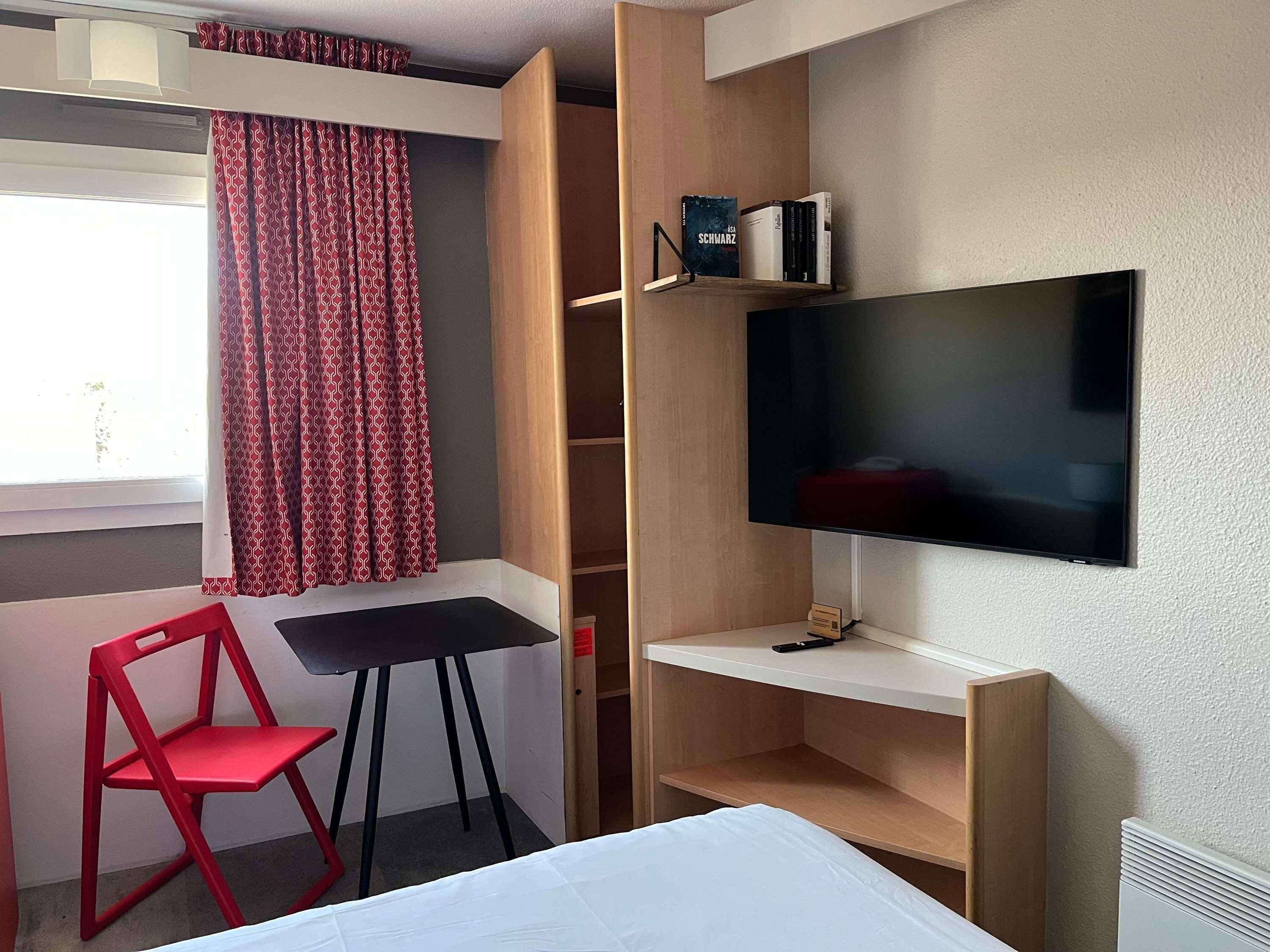 Une chambre d’hôtel avec un lit, un bureau, une télévision, une chaise rouge et une fenêtre avec des rideaux à motifs.