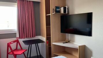 A hotel room with a bed, a desk, a TV, a red chair, and a window with patterned curtains.