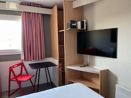Habitación de hotel con cama, escritorio, televisor, una silla roja y ventana con cortinas estampadas.