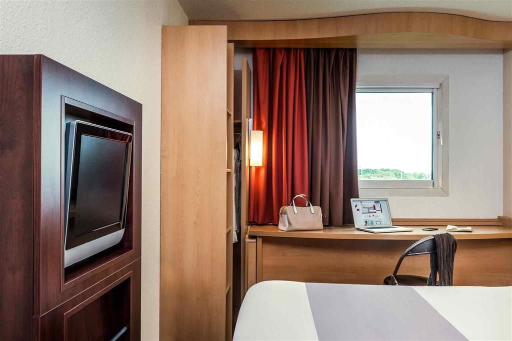 A hotel room with a TV, a desk with a laptop, a chair, and a window with curtains.